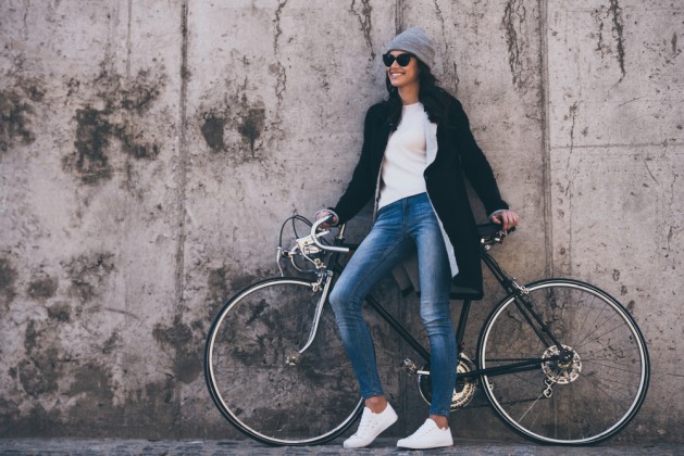 That was great ride! Beautiful young woman in sunglasses holding hands on her bicycle and looking away with smile while standing against concrete wall outdoors