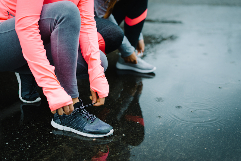 Urban athletes lacing sport footwear for running over asphalt under the rain. Two women getting ready for outdoor training and fitness exercising on cold winter weather.
