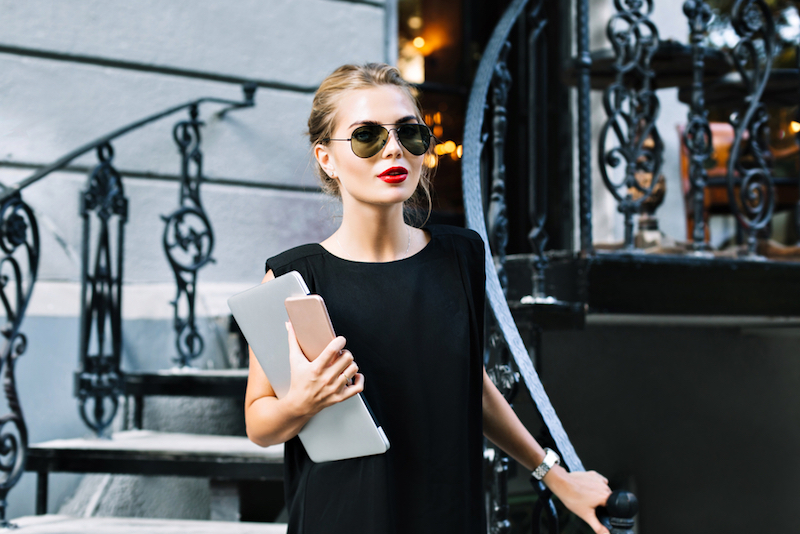 Portrait pretty businesswoman in black dress on stairs outdoor. She wears sunglasses, laptop, phone, looking to camera