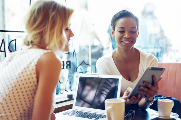 Two entrepreneurs are smiling while discussing news displayed on a screen of a digital tablet. International female students are working on a new project by using portable computer and a tablet eating lunch at work