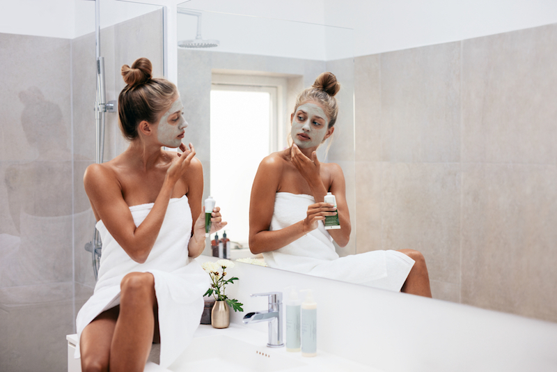 Young woman applying facial mud clay mask to her face in bathroom. Beautiful female wrapped in towel looking into mirror and doing facial beauty treatment.