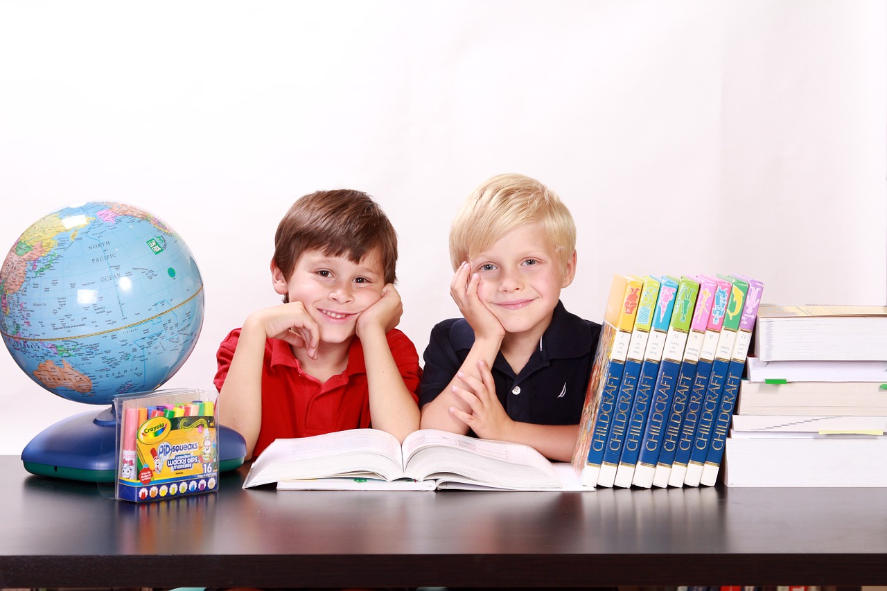 Two children in classroom setting reading a book, next to globe and crayons
